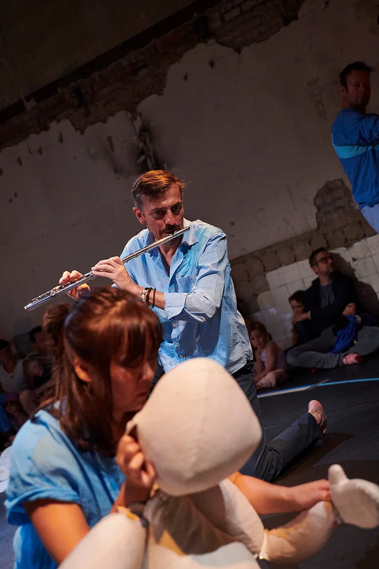 Ein Flötenspieler trägt ein hellblaues Hemd, spielt Flöte und beobachtet eine Schauspielerin, die mit einer Handpuppe spielt