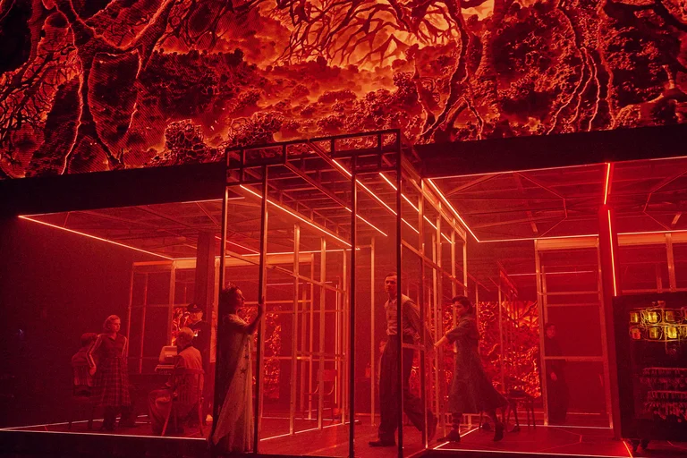 Stage scene in red lighting with several actors inside a metal structure, above them a projection of organic-looking patterns.