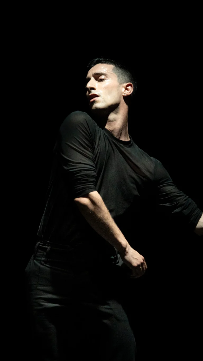 Performer Georgios Kotsifakis in dark clothes moving vigorously in front of a dark background.