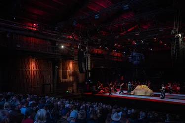 The audience watches a performance in a dark industrial hall. On the red-lit stage, several performers move around a large fabric-covered object while musicians play at the side of the stage.