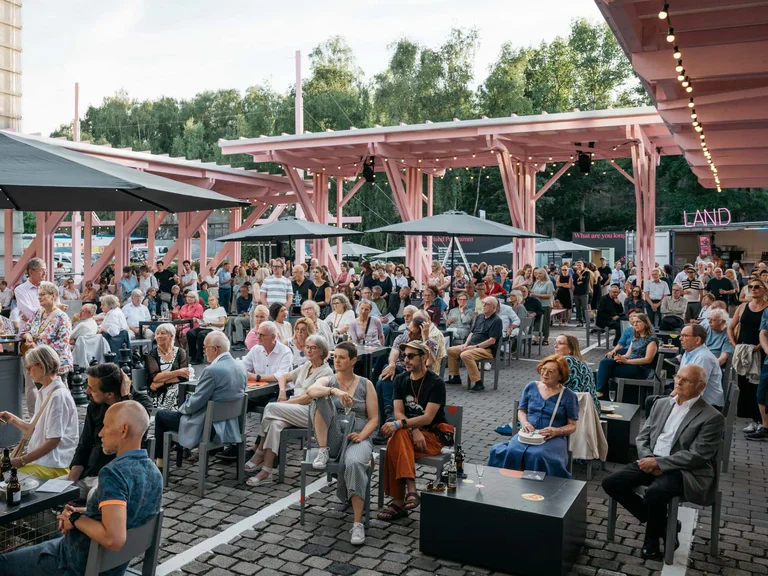 A lively audience under the pink wooden structure which is the festival centre Wunderland, with event booths visible in the background.