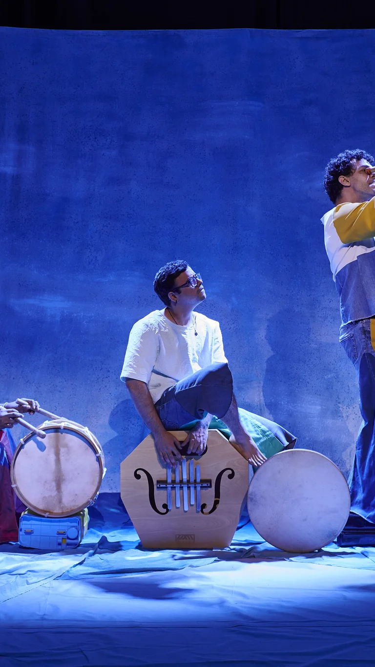 Drei Musiker auf einer blau beleuchteten Bühne spielen Trommeln und ein langes Blasinstrument.