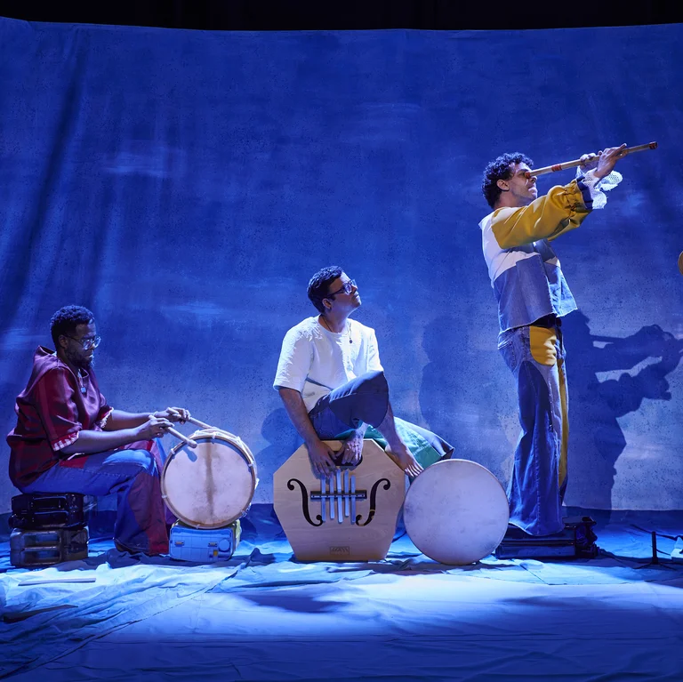 Three musicians on a blue-lit stage play drums and a long wind instrument.