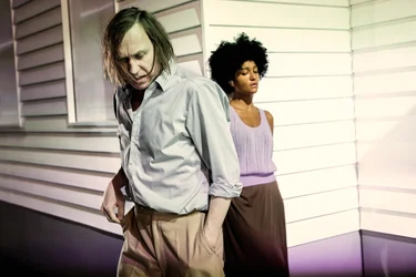 Larissa Sirah Herden stands calmly with crossed arms in the background, while Lars Eidinger walks past her in the foreground with a downward gaze — the scene conveys tension.