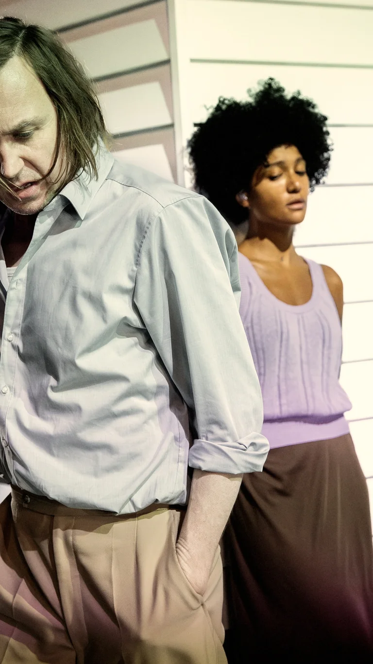 Larissa Sirah Herden stands calmly with crossed arms in the background, while Lars Eidinger walks past her in the foreground with a downward gaze — the scene conveys tension.