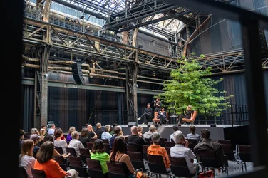 Blick auf die Bühne bei der Diskursveranstaltung "Brave New Voices" mit Autorin und Dramatikerin Sivan Ben Yshai. Im Vordergrund sieht man das Publikum, im Hintergrund die Bühne mit den Akteur:innen im Gespräch.