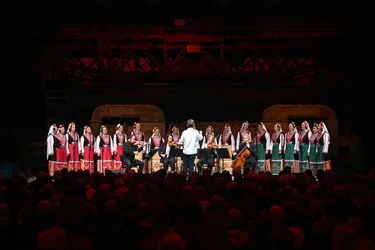 Ein großer Frauenchor in traditionellen, farbenfrohen Trachten singt auf einer Bühne, begleitet von einem Streichensemble. Ein Dirigent steht vor den Musiker:innen und Sängerinnen, während ein großes Publikum die Aufführung verfolgt