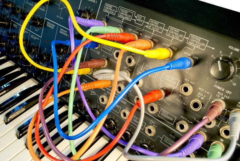Ein analoger Synthesizer mit zahlreichen bunten Patchkabeln, die in verschiedene Buchsen eingesteckt sind. Die Oberfläche des Synthesizers zeigt komplexe Beschriftungen und Bedienelemente wie Regler und Schalter. Im unteren Bereich sind Tasten einer Klaviatur sichtbar.