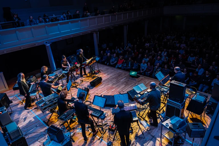 Das Will Gregory Moog Ensemble bei einer Live-Performance auf einer Bühne in einem Konzertsaal. Mehrere Musiker stehen oder sitzen in einem Halbkreis, umgeben von Synthesizern, Notenständern und elektronischen Instrumenten. Das Publikum, das im Hintergrund zu sehen ist, sitzt in einem gedämpften blauen Licht.