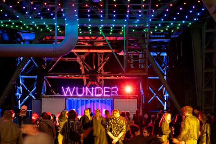 A crowd of visitors celebrates under colourful lights in front of the illuminated WUNDER sign at the festival centre.