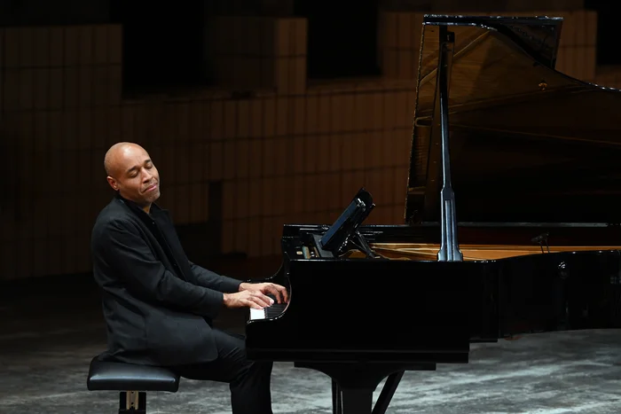 Pianist Aaron Diehl sits at the grand piano, playing with his eyes closed. He is wearing a black jacket and appears deeply immersed in the music.