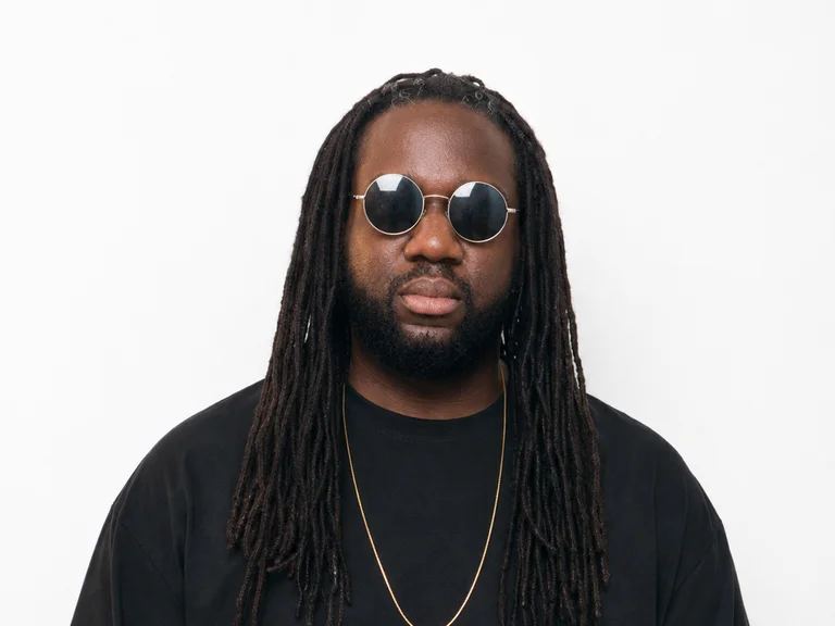 DJ Spread Out steht, Sonnenbrille und ein schwarzes T-Shirt tragend, vor einer hellen Wand.