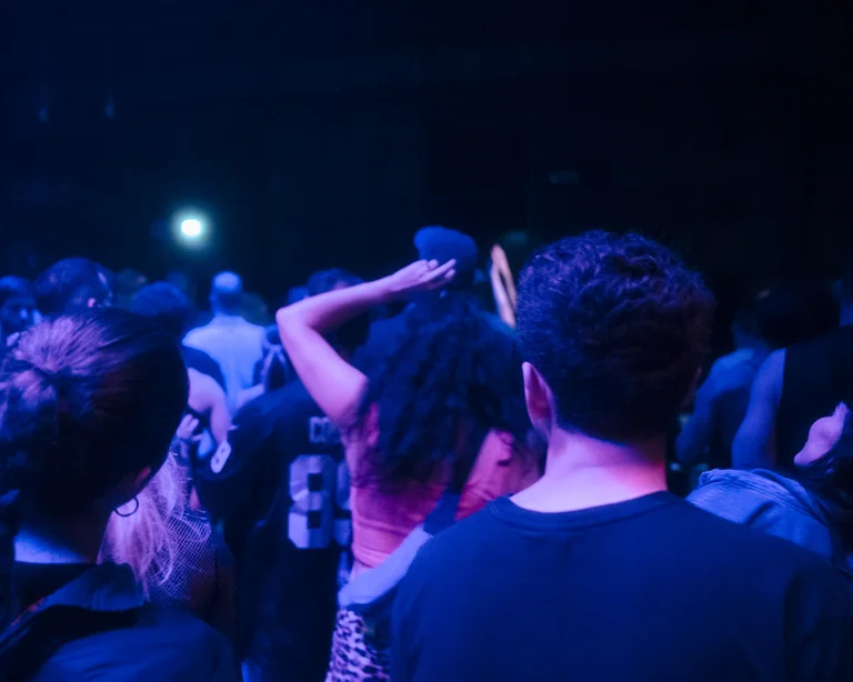 People stand closely packed in a dark hall, bathed in blue-violet light. A woman in a red top raises her arm as the crowd moves within the club atmosphere.
