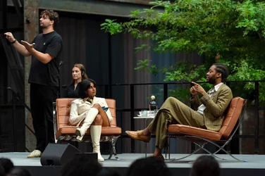 Das Bild zeigt eine Nahaufnahme der Gesprächsrunde im Rahmen der Veranstaltung „Brave New Voices“ in der Jahrhunderthalle Bochum, Teil der Ruhrtriennale. Auf der Bühne sitzt die Moderatorin Fatima Khan in einem weißen Outfit, während sie dem Schriftsteller Jeremy O. Harris zuhört, der in einem gestreiften Anzug spricht. Im Hintergrund steht ein Gebärdensprachdolmetscher, der die Unterhaltung übersetzt. Die Bühne ist mit Bäumen dekoriert, und man erkennt die industrielle Architektur der Halle.