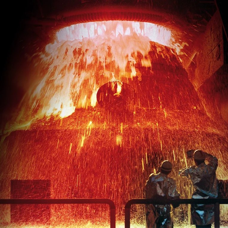 Zwei Arbeiter in Schutzkleidung stehen vor einem Hochofen, aus dem glühendes, flüssiges Metall in einem intensiven Funkenregen herabströmt.