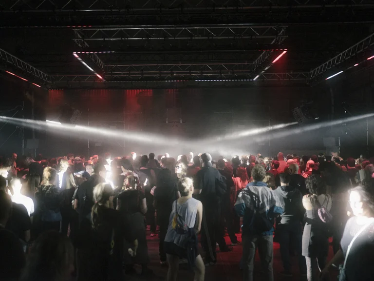 A large crowd dances in a hall as bright beams of light and red stage lighting illuminate the scene dramatically.