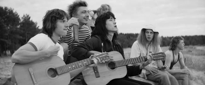 Schwarz-weiß Film still aus dem Film "Leto", auf dem mehrere Personen am Strand sitzen und Gitarre spielen.