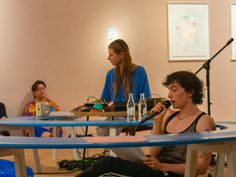 A person sits on the floor behind a table, reading from a text into a microphone, while another person operates audio equipment at a table. Another person sits in the background on the floor.