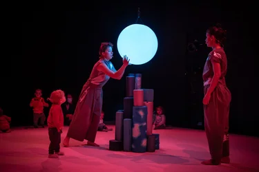 Szene aus der partizipativen Performance UNIVERS: Eine Performerin berührt eine schwebende, leuchtende Kugel, während Kinder aus nächster Nähe zusehen. Die Atmosphäre ist ruhig und konzentriert.