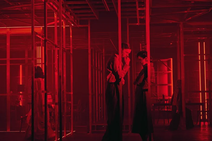Actors in a stage structure of metal frames illuminated by red lights.