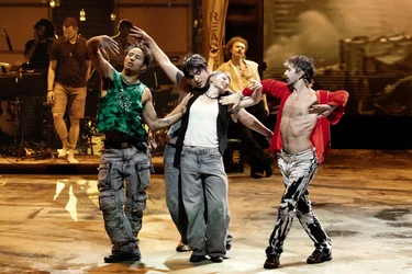 Several performers move across the stage in a group choreography. In the foreground, four people stand close together with their arms outstretched and intertwined. In the background, other performers and a band can be seen. The scene is bathed in warm, golden light.