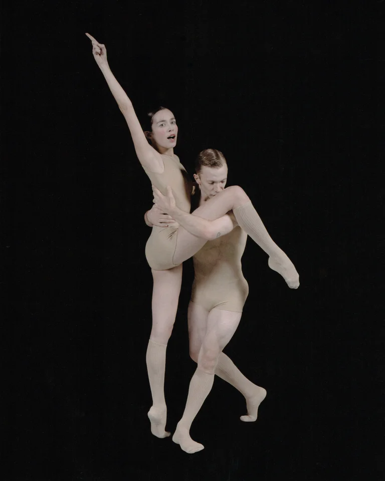 A dancer lifts a partner in a skin-toned costume as she stretches one arm expressively upward. The pose combines strength and lightness, creating a sense of elevation.