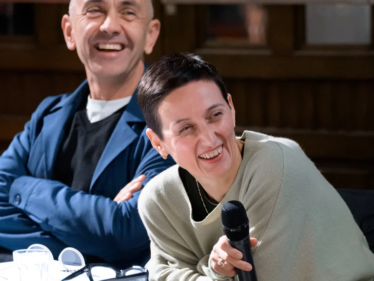 Ruth Anderwald und Leo Grond, die nebeneinander an einem Tisch sitzen und sich lachend zur Seite drehen. Ruth Anderwald, vorne rechts im Bild, hält ein Mikrofon in den Händen.