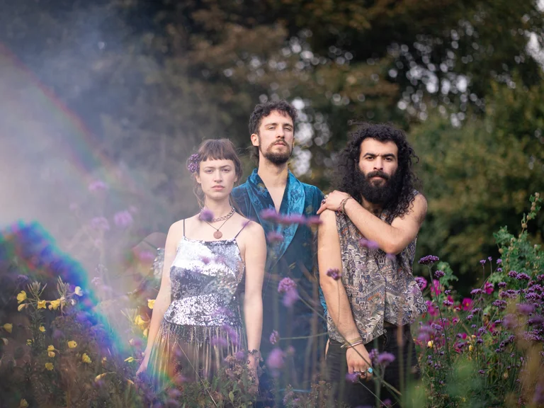 The three band members of Karmakind stand in a blooming garden surrounded by purple flowers. A soft rainbow shimmers in the image.