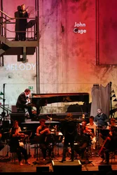 The ensemble Les Apaches ! performs on stage in an industrial hall. At the centre, Julien Masmondet conducts the musicians while a pianist plays the grand piano. Another musician stands on a metal staircase holding mallets. The stage and the wall in the background are bathed in red light, with the name “John Cage” projected onto the wall.