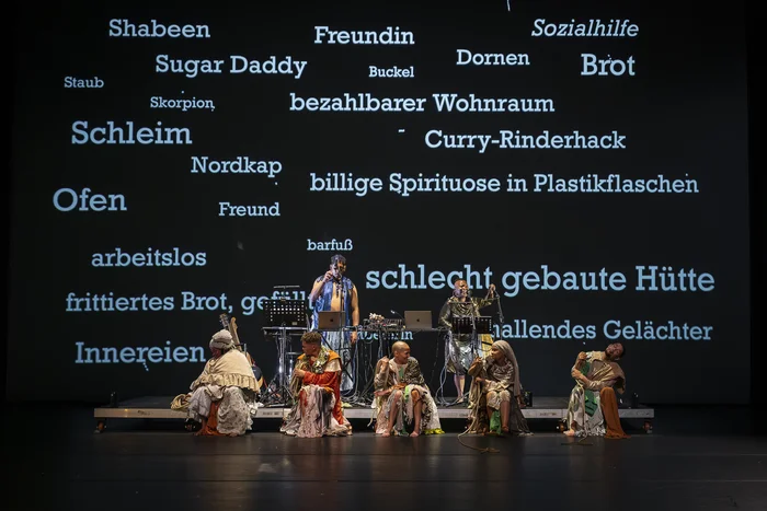 Eine Bühnenperformance mit fünf Darsteller:innen in bunten Gewändern, im Hintergrund werden deutsche Wörter und Begriffe groß auf eine Leinwand projiziert.
