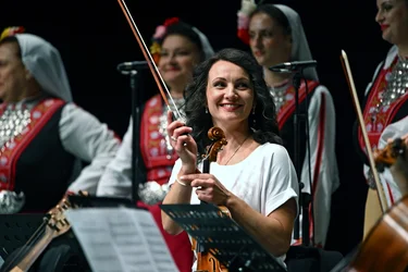 Eine lächelnde Geigerin sitzt mit ihrem Instrument und Bogen auf der Bühne. Im Hintergrund sind Sängerinnen in farbenfrohen traditionellen Trachten zu sehen.