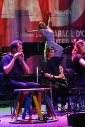 Sarah Silverblatt-Buser dances with her arms stretched out wide behind the musicians of the ensemble Les Apaches !. In the foreground, two musicians play wind instruments. The stage and the projections on the back wall are bathed in red, purple and green light.