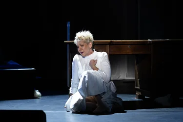 Joyce DiDonato sits barefoot in a white dress on a dimly lit stage in front of a wooden table. One hand rests on her chest as she sings with her eyes closed.