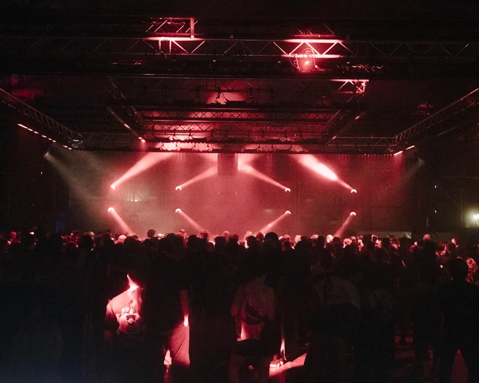 A large crowd stands in a hall in front of a stage, with red spotlights shining through the room.