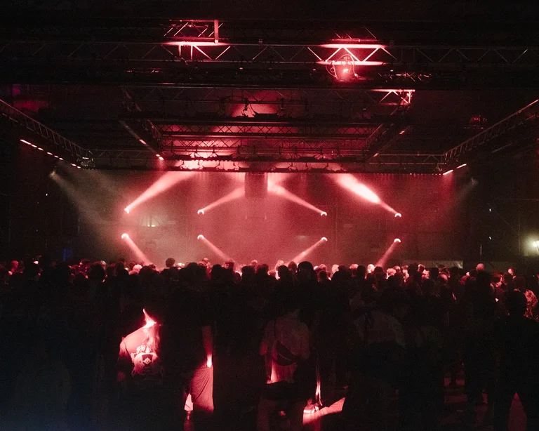 A large crowd stands in a hall in front of a stage, with red spotlights shining through the room.