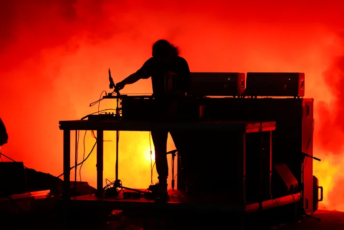 James Ginzburg stands behind a table with electronic equipment, his figure silhouetted against intense red fog. Cables and technical devices are outlined in the backlight.