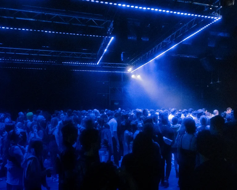 A large crowd dances closely together under blue lighting in a club-like hall. The geometric lighting from above enhances the energetic and atmospheric mood.