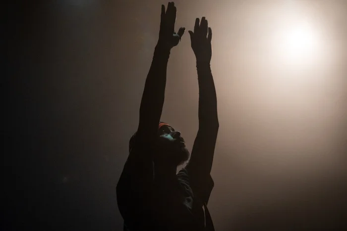 Silhouette of Mohamed Toukabri with arms raised against backlight, light cutting through stage haze from above.