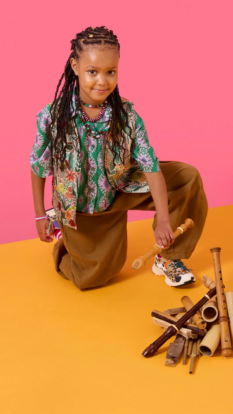 Ein Kind kniet auf dem Boden und hält Holzklangstäbe in der Hand; neben ihm liegen weitere Klangobjekte und eine Trommel.