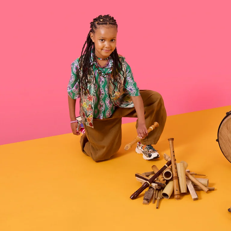 Ein Kind kniet auf dem Boden und hält Holzklangstäbe in der Hand; neben ihm liegen weitere Klangobjekte und eine Trommel.