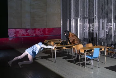 Eine Person liegt mit ausgestrecktem Körper über einem Tisch, während eine zweite Person auf der gegenüberliegenden Seite des Tisches steht. Mehrere Schulbänke strukturieren den Bühnenraum.