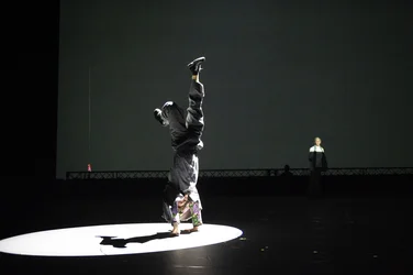 Solal Mariotte balanciert im Handstand auf einer runden, beleuchteten Fläche; im Hintergrund steht Anne Teresa De Keersmaeker im Halbdunkel.