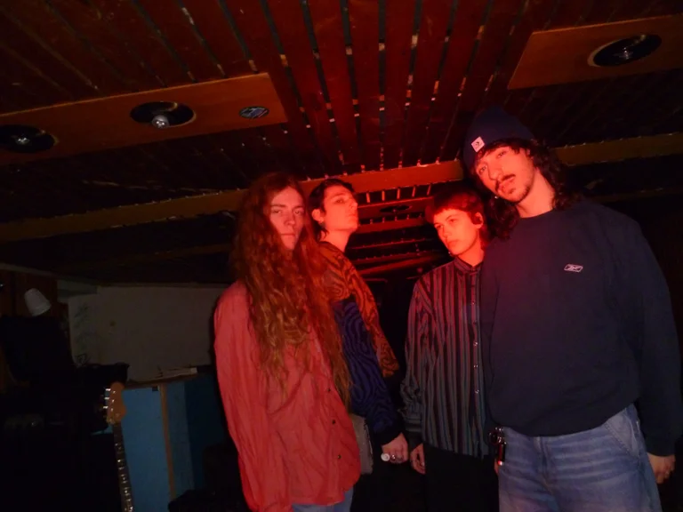 The four band members of The Destruents stand under red lighting in a rehearsal room with a wooden ceiling, looking toward the camera.