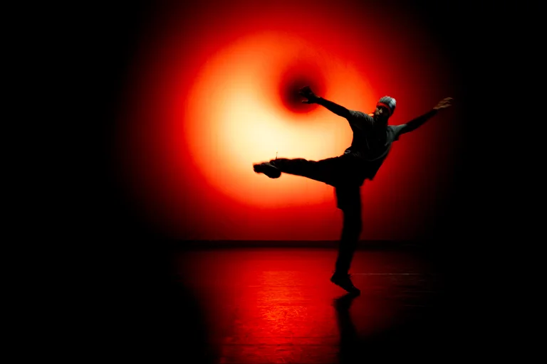 Mohamed Toukabri tanzt dynamisch vor einem leuchtend roten Lichtkreis auf dunkler Bühne. Seine Silhouette ist nur schemenhaft erkennbar und steht im starken Kontrast zum hellen Hintergrund.