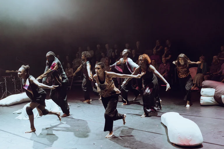 A group of seven dancers moves together away from the audience, who are seated on stage in the background. White sacks lie on the dark stage.