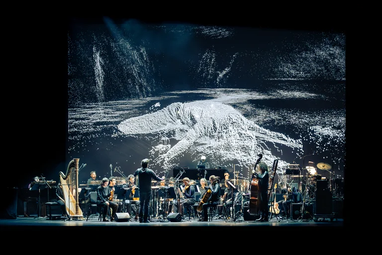 Les Apaches! is performing on a stage with a large digital backdrop. Julien Masmondet stands in the foreground with his back to the audience, while musicians with string and wind instruments, as well as a harp and percussion, form the ensemble.