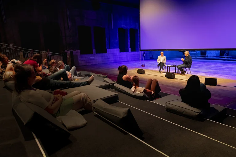 Audience in an industrial hall watching a conversation between Ivo Van Hove and Will Gregory on stage in front of a large screen.