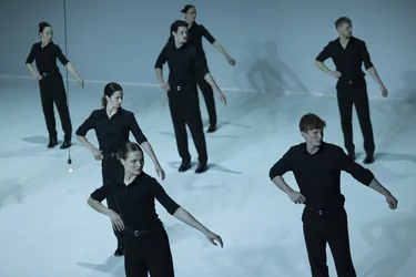 Several performers in black clothing move with extended arms on a bright stage.