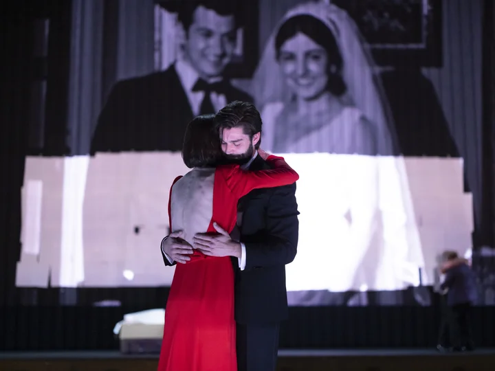 A nostalgic film scene with a wedding couple is projected in the background, while a couple on stage loses themselves in an embrace.