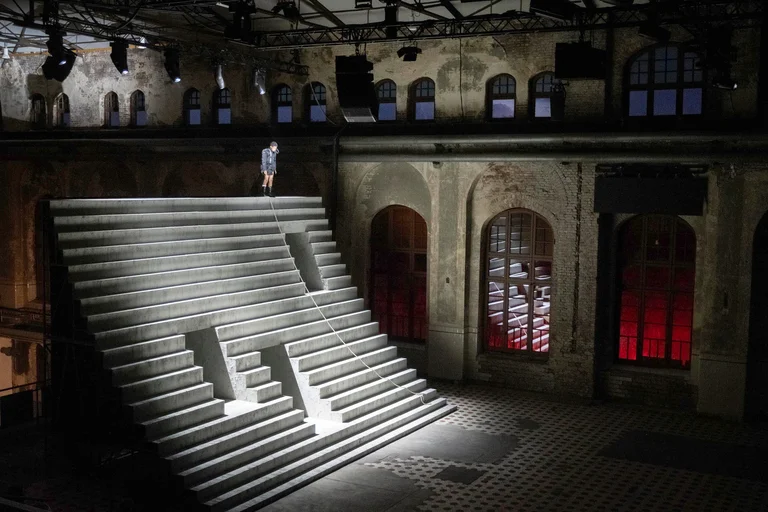 Eine einzelne Person steht oben auf einer monumentalen, hell beleuchteten Treppenkonstruktion in einer weitläufigen Industriehalle. Ein langes Seil führt von der Person über die Stufen bis zum Boden.
